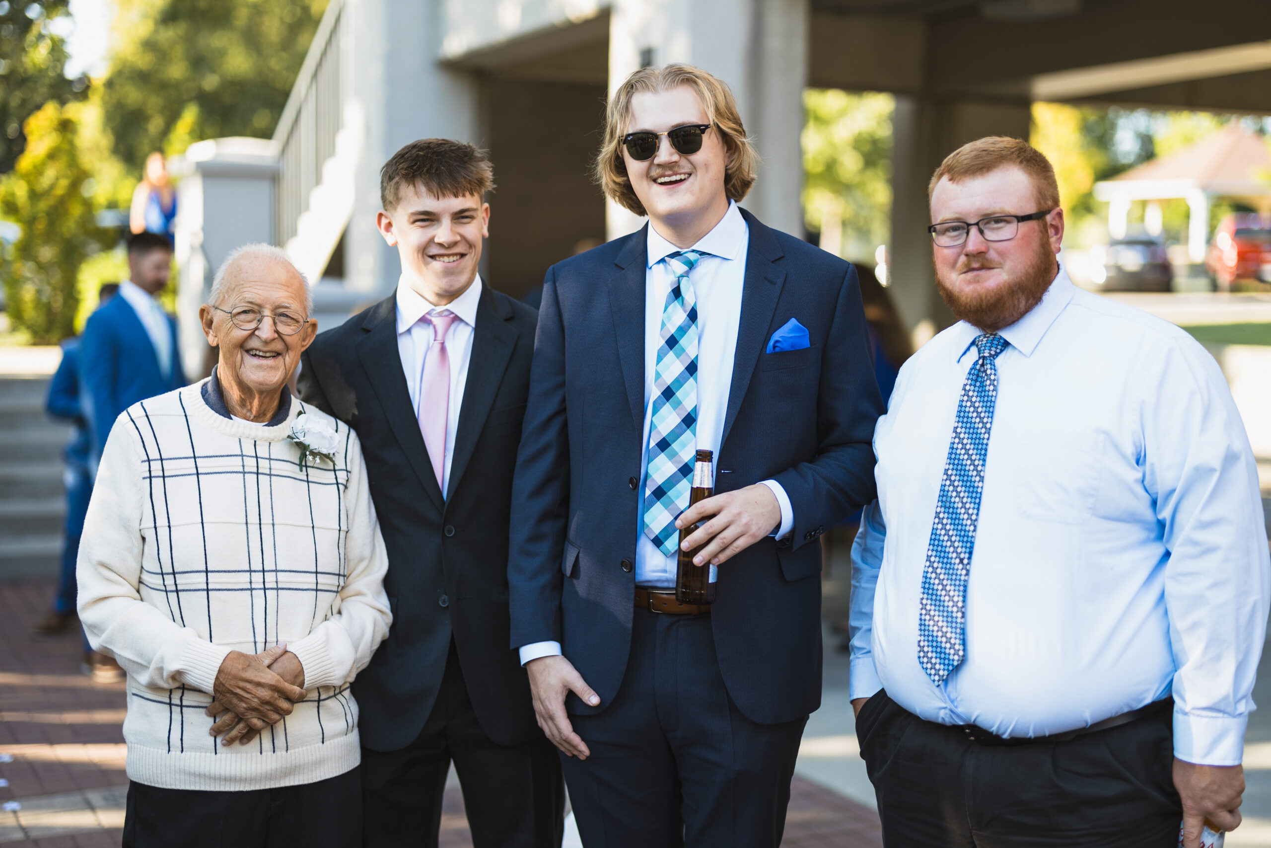 Group of smiling people in formal attire posing outdoors at a sunny event, including an elderly woman in a cream sweater and three men in suits.