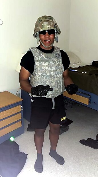 Person wearing camouflage helmet, tactical vest, gloves, and black clothing, posing with peace signs in a dorm room.