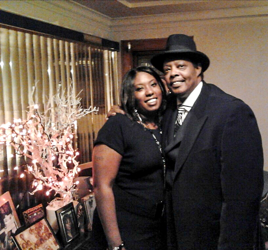 A smiling couple posing together indoors, man in a dark suit and fedora beside a woman in a black dress, with a decorative lit tree and photos on a table nearby.