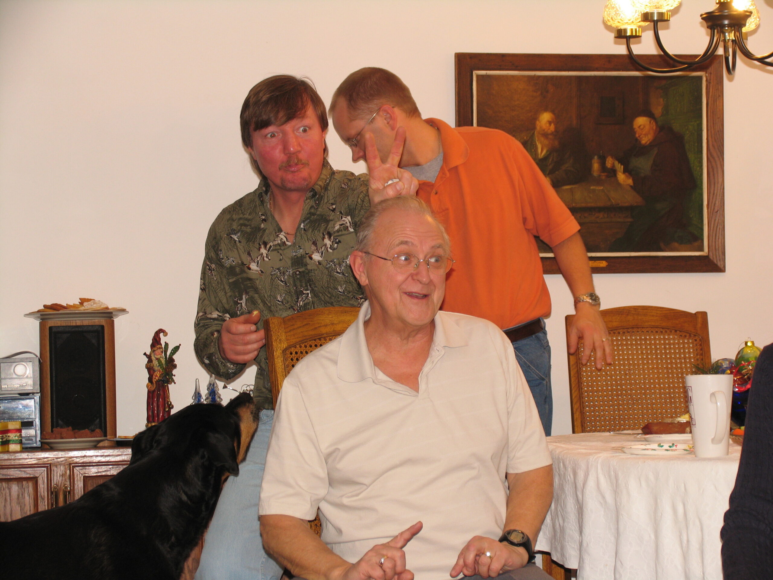 Older man with glasses seated at a table; a younger man in an orange shirt stands behind him, making a peace sign near his head. A black dog sits in the foreground, with a plate of cookies on the left and a painting on the wall in the background.