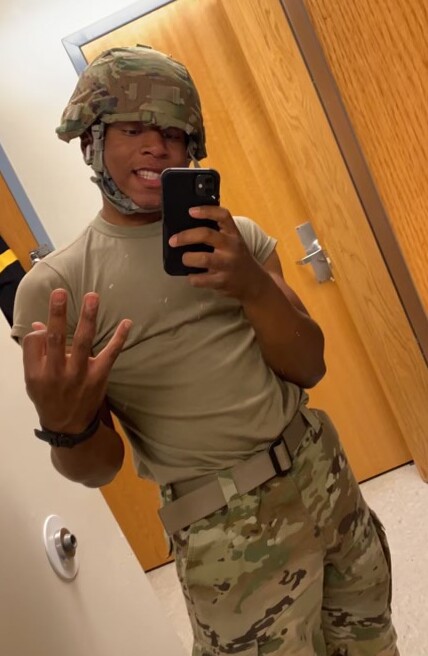 Person in desert camouflage uniform and helmet taking a mirror selfie with a smartphone, making a three-finger gesture.