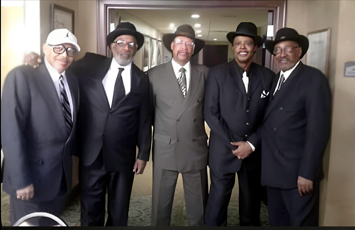 Five men in dark suits and hats pose together in a hallway, smiling for the camera.