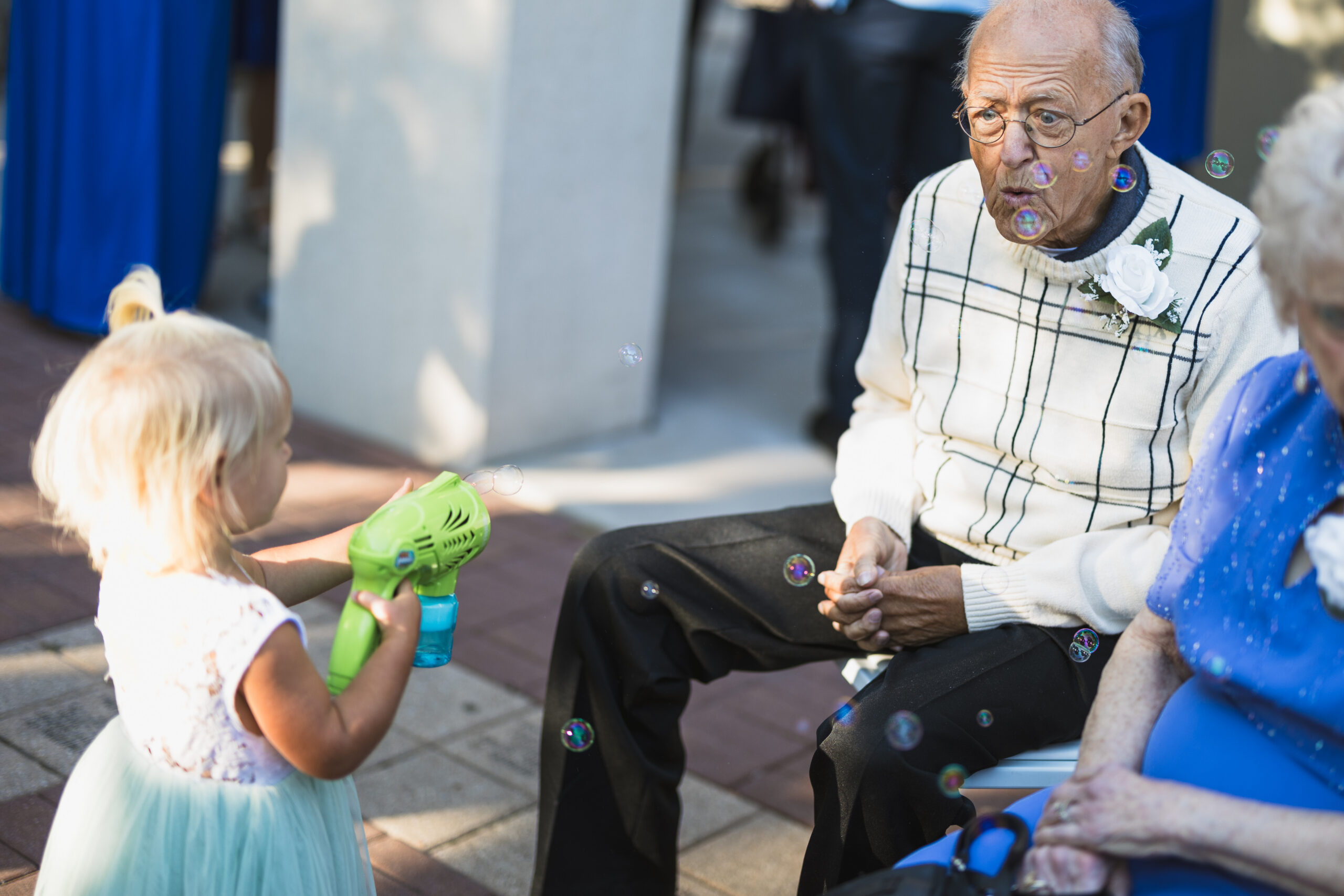 Child in a light dress hands a green bubble blower to an elderly man as bubbles float around them outdoors.
