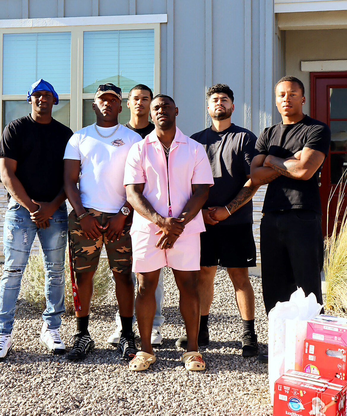 Six men stand in a line outside a house, posing for a group photo with gift boxes nearby.