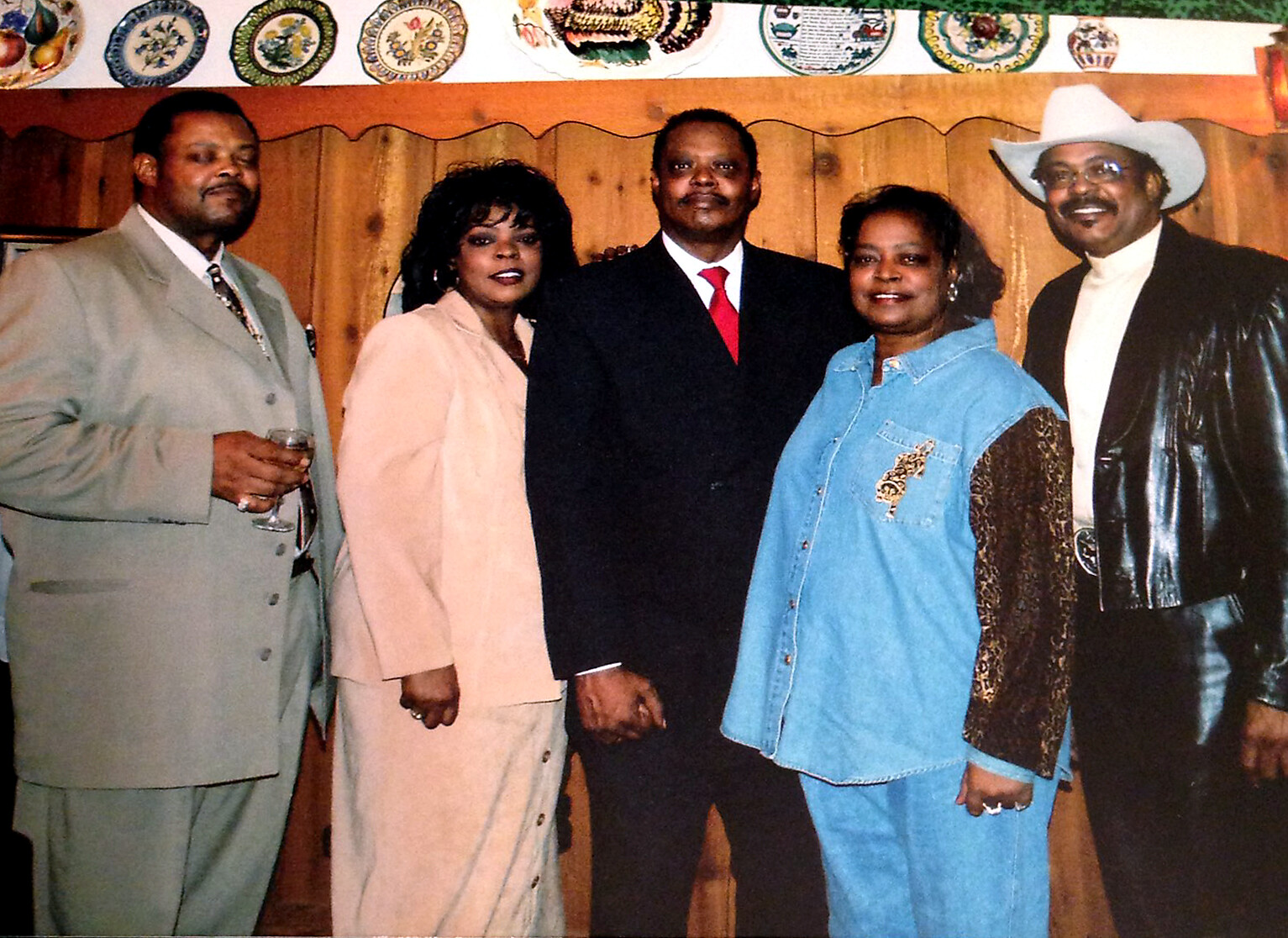 Five adults posing indoors against wood paneling; two men in suits, two women, and a man wearing a white cowboy hat.