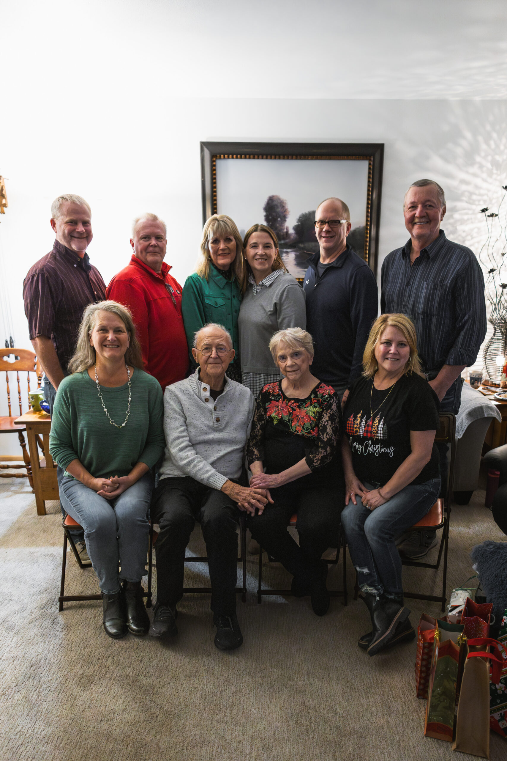 Ten adults posing for a group photo in a living room, smiling, with framed art on the wall and gift bags on the floor to the right.