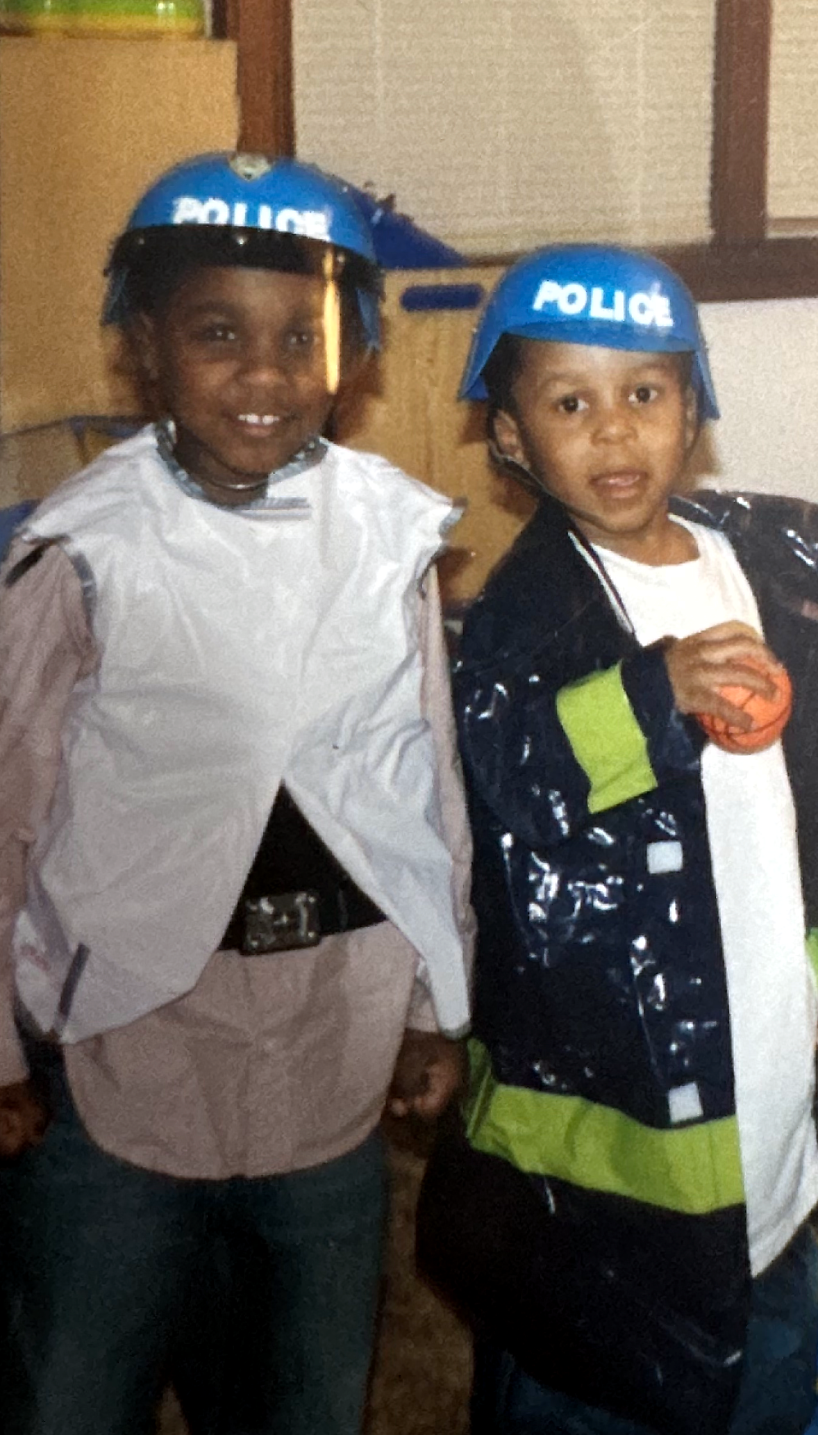 Two children wearing blue toy police helmets pose indoors; one in a gray cape and belt, the other in a black jacket holding an orange ball and looking at the camera.