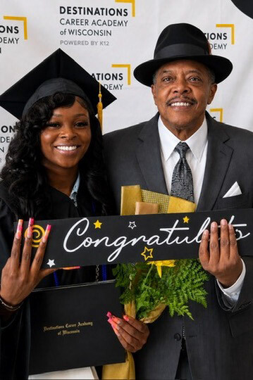 Graduate in cap and gown beside a man in a suit, holding a 'Congratulations' banner and a bouquet against a ceremony backdrop by DESTINATIONS ACADEMY