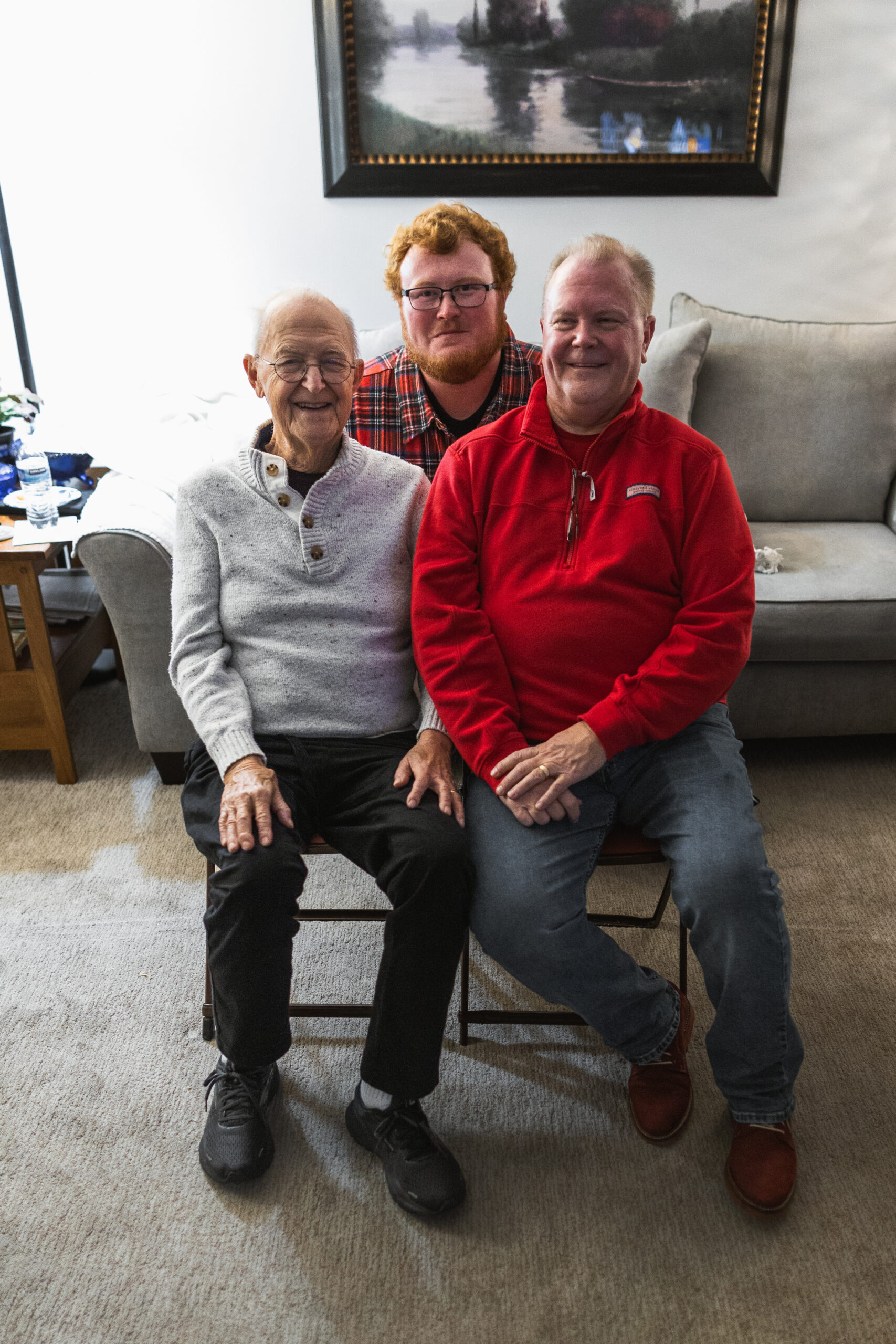 Three men of different ages sit close together on a beige sofa, smiling at the camera in a living room.