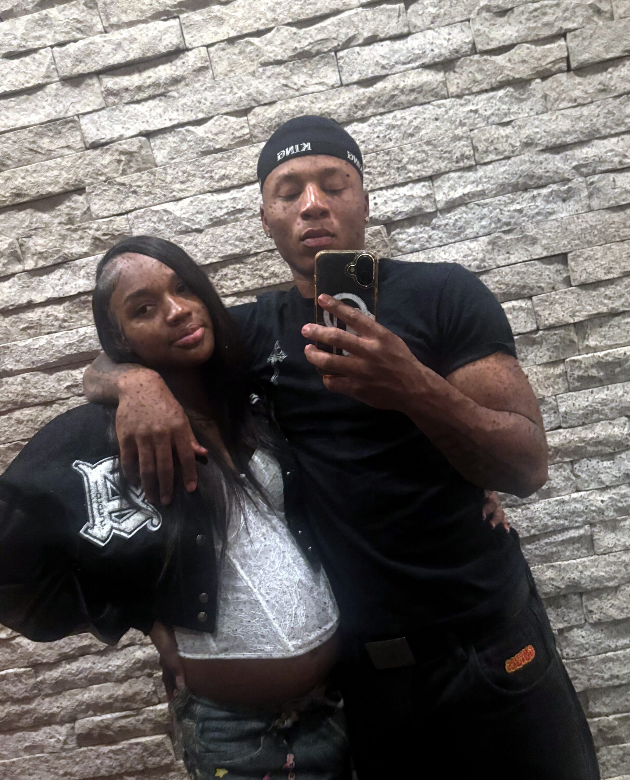 Couple posing for a selfie in front of a textured stone wall; man in a black shirt and headband, woman in a dark jacket beside him, both smiling slightly.