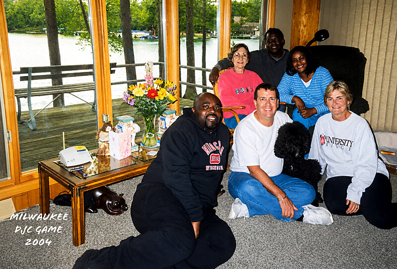 Group photo of diverse adults and a black dog in a living room by a lake, with flowers on a side table and 'MILWAUKEE PJC GAME 2004' text on the image.