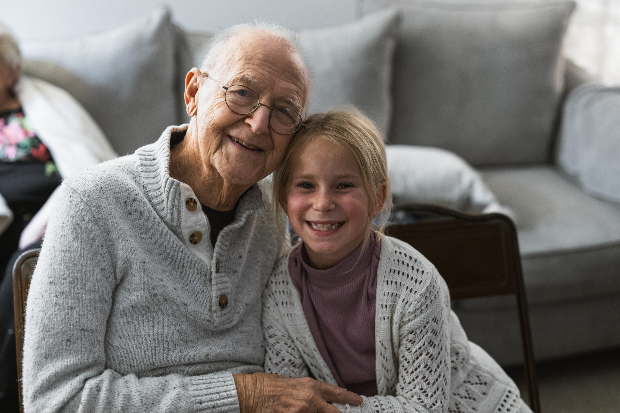 Grandmother and young girl smiling together on a couch, arms around each other in a cozy living room setting.