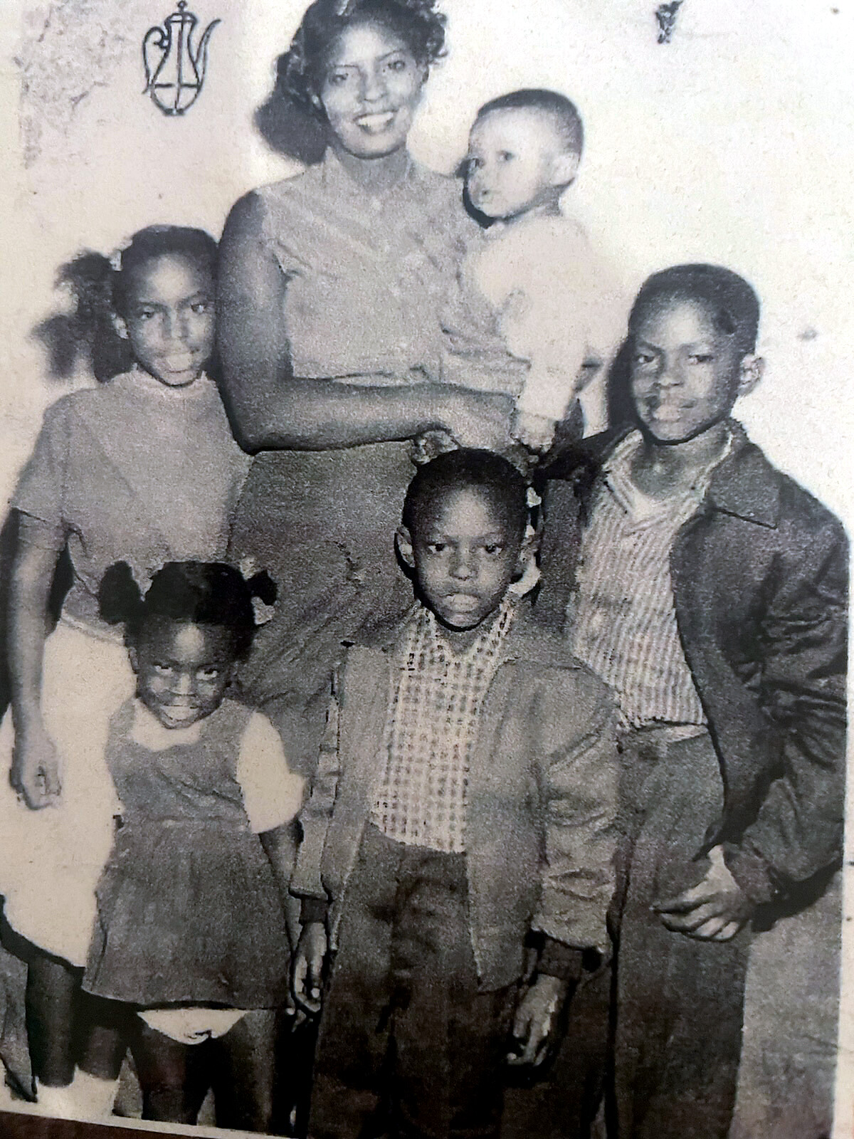 A mother holds a baby while five children stand around her in a black-and-white family photo from the past.