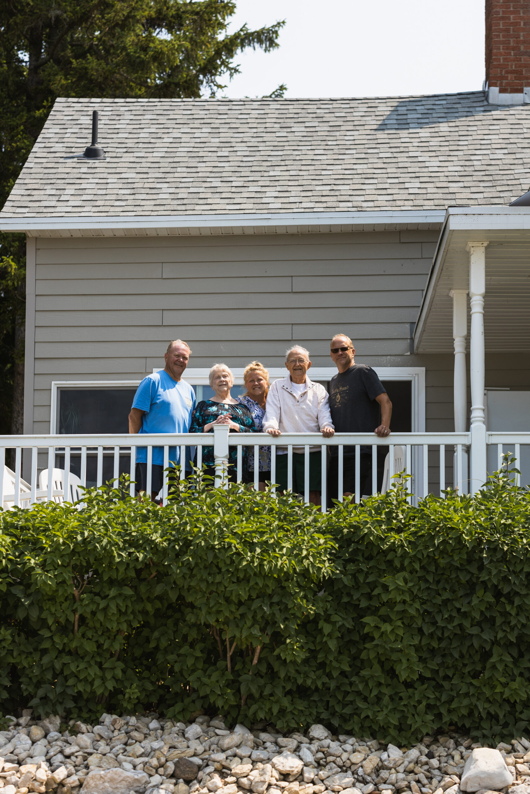 Five adults smiling on a white railing balcony in front of a light gray house, with a tall green hedge in the foreground.