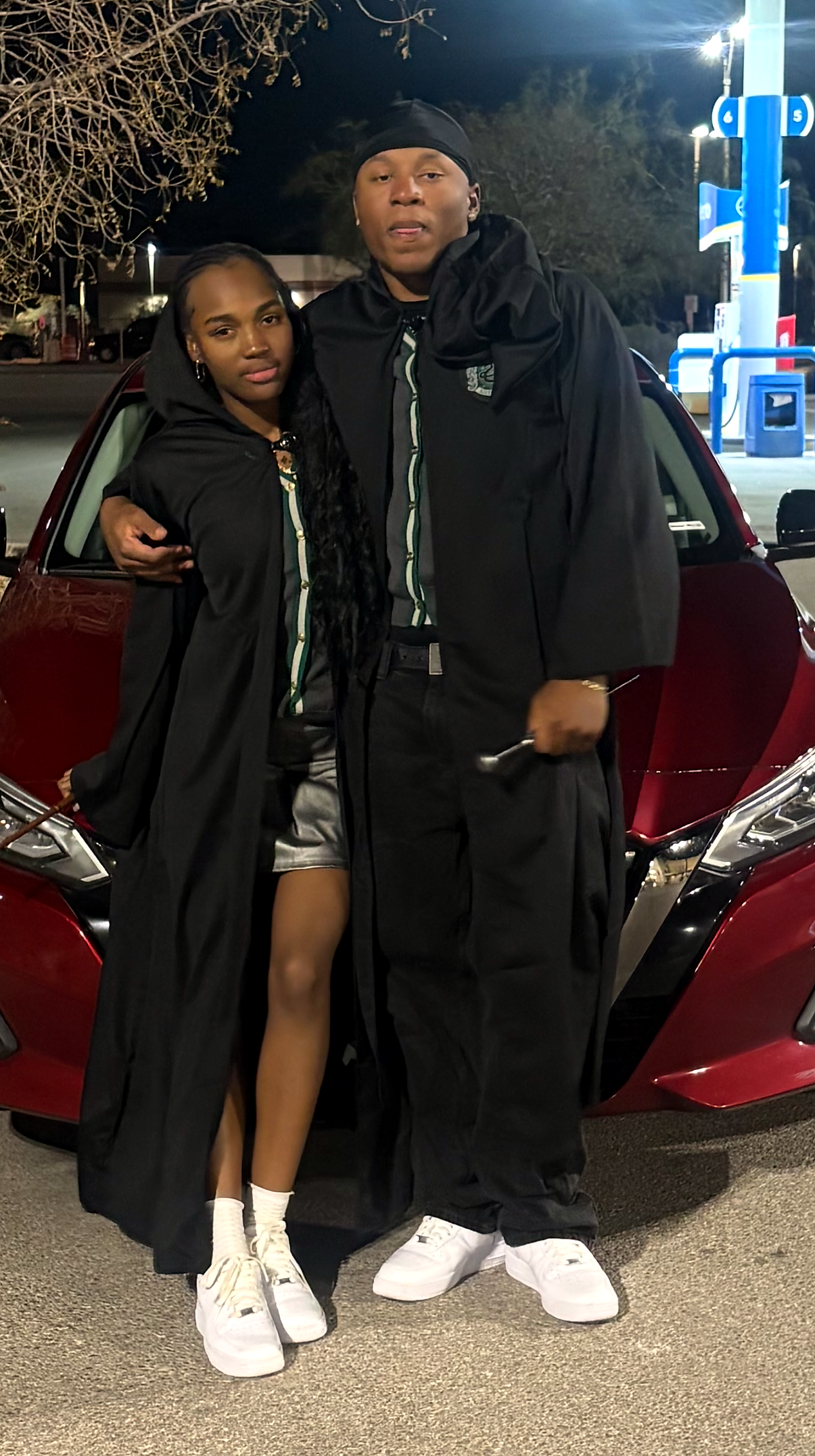 Two people in black graduation robes pose together in front of a red car at night.