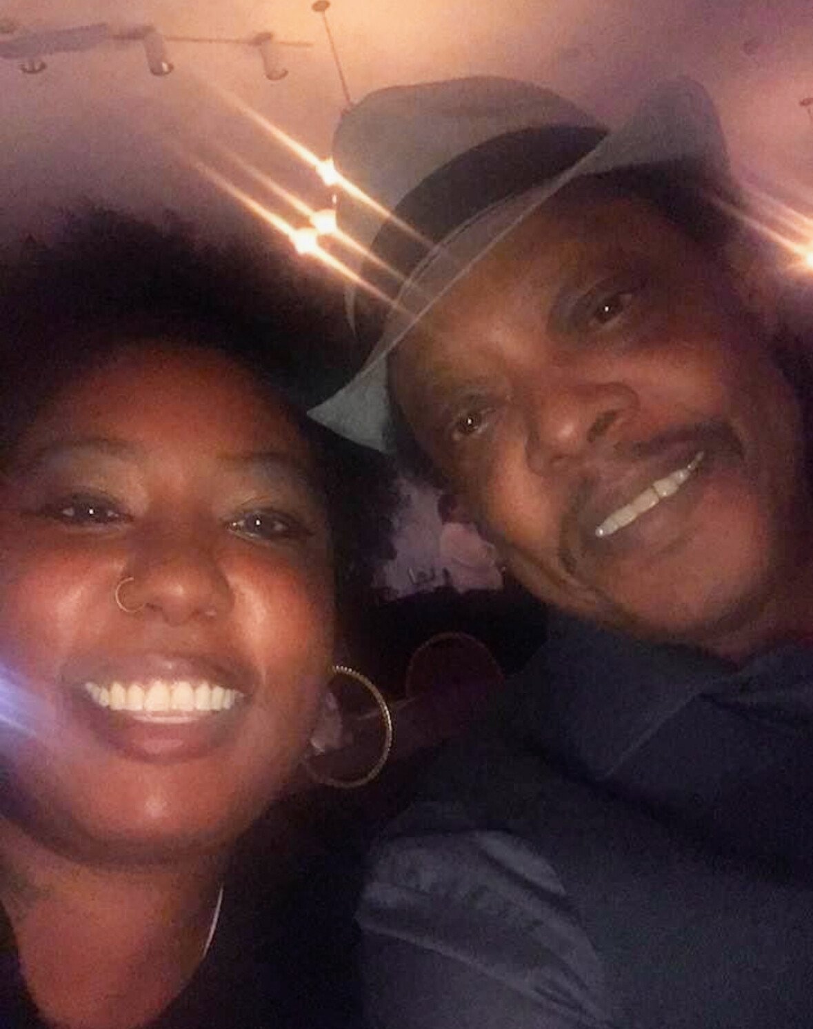 Two people smiling for a selfie in a dimly lit venue with warm string lights overhead.