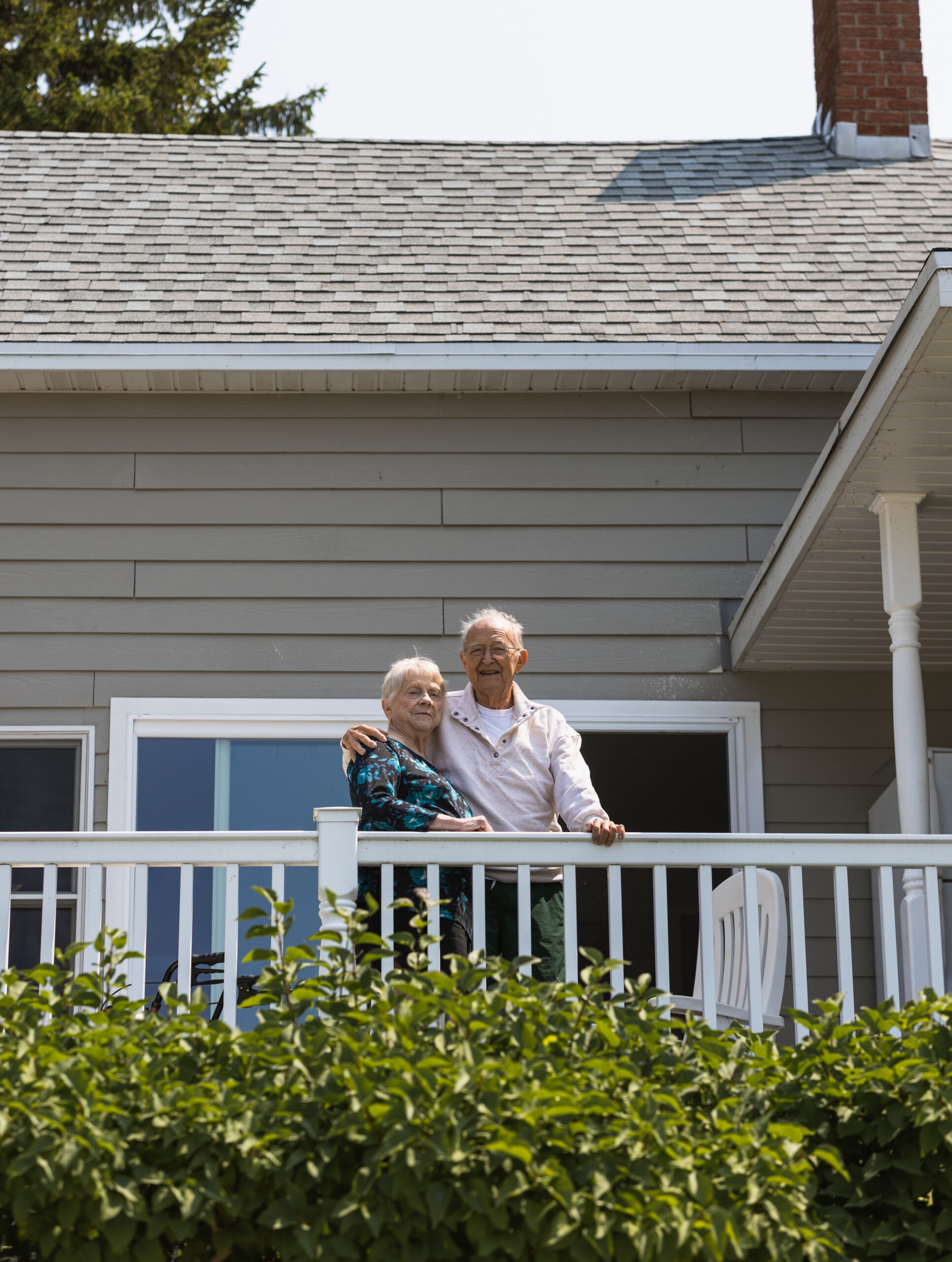 Two elderly people stand on a white balcony railing outside a gray siding house, smiling, with green bushes in the foreground.