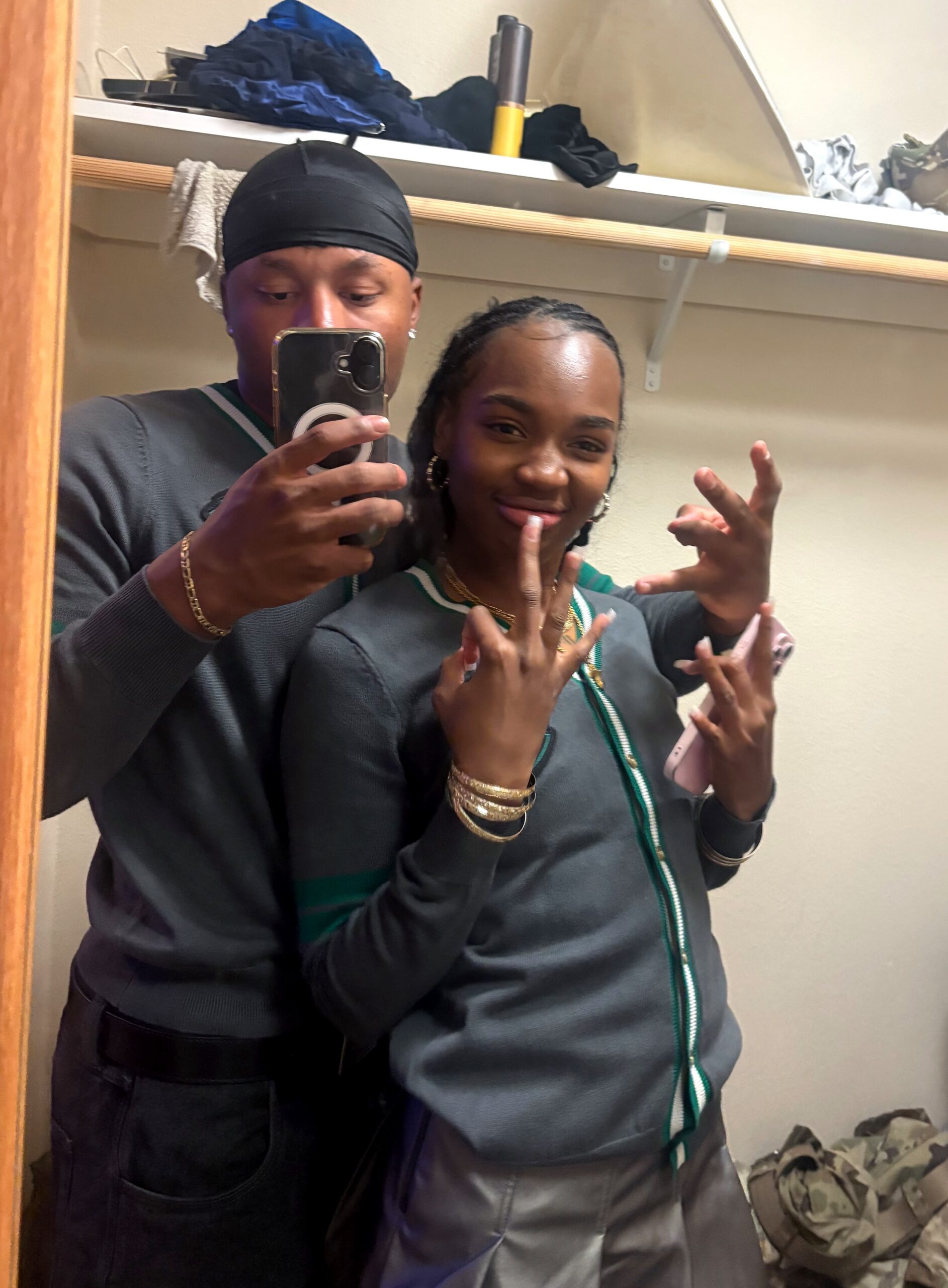 Two friends take a mirror selfie in a cluttered room wearing gray jackets with green trim; the man wears a black durag and gold bracelets while the woman smiles and poses with hand gestures.