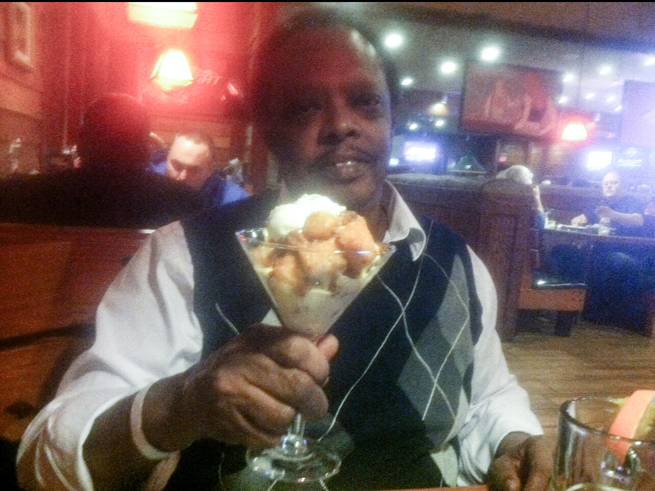 Man in a restaurant holding a tall glass dessert with scoops of ice cream and whipped cream, looking at the camera.