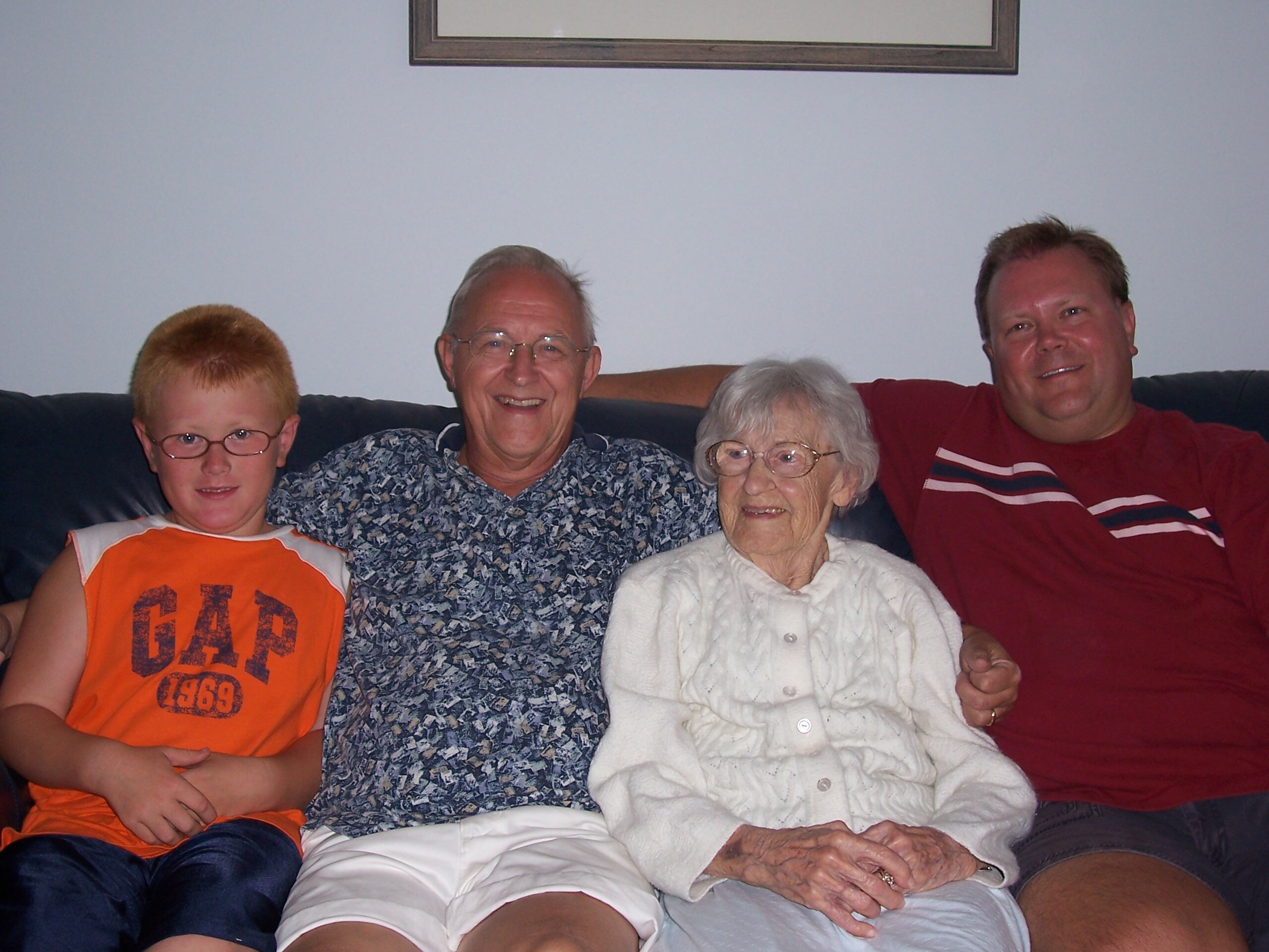 Four people sitting on a sofa smiling at the camera: a boy in an orange shirt, two older adults, and a man in a red shirt.
