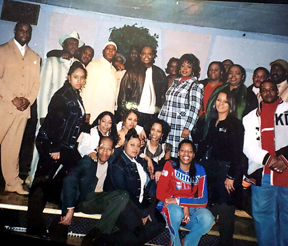 Large group portrait of about 20 people posing on steps indoors, wearing varied outfits from denim jackets to suits, with many smiling at the camera.