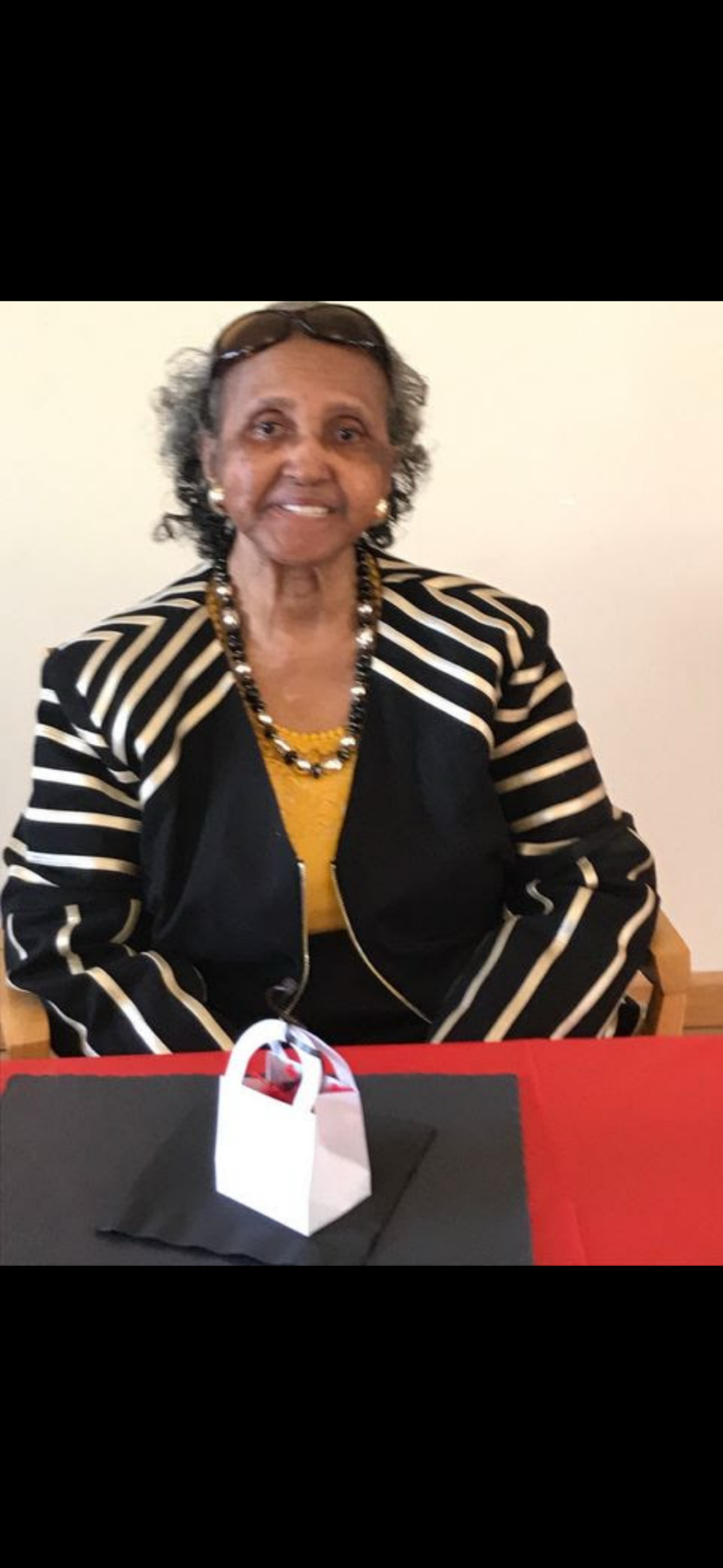 Smiling elderly woman in a black‑and‑white striped jacket with a yellow top, sitting at a table with a small white gift bag on a black napkin.
