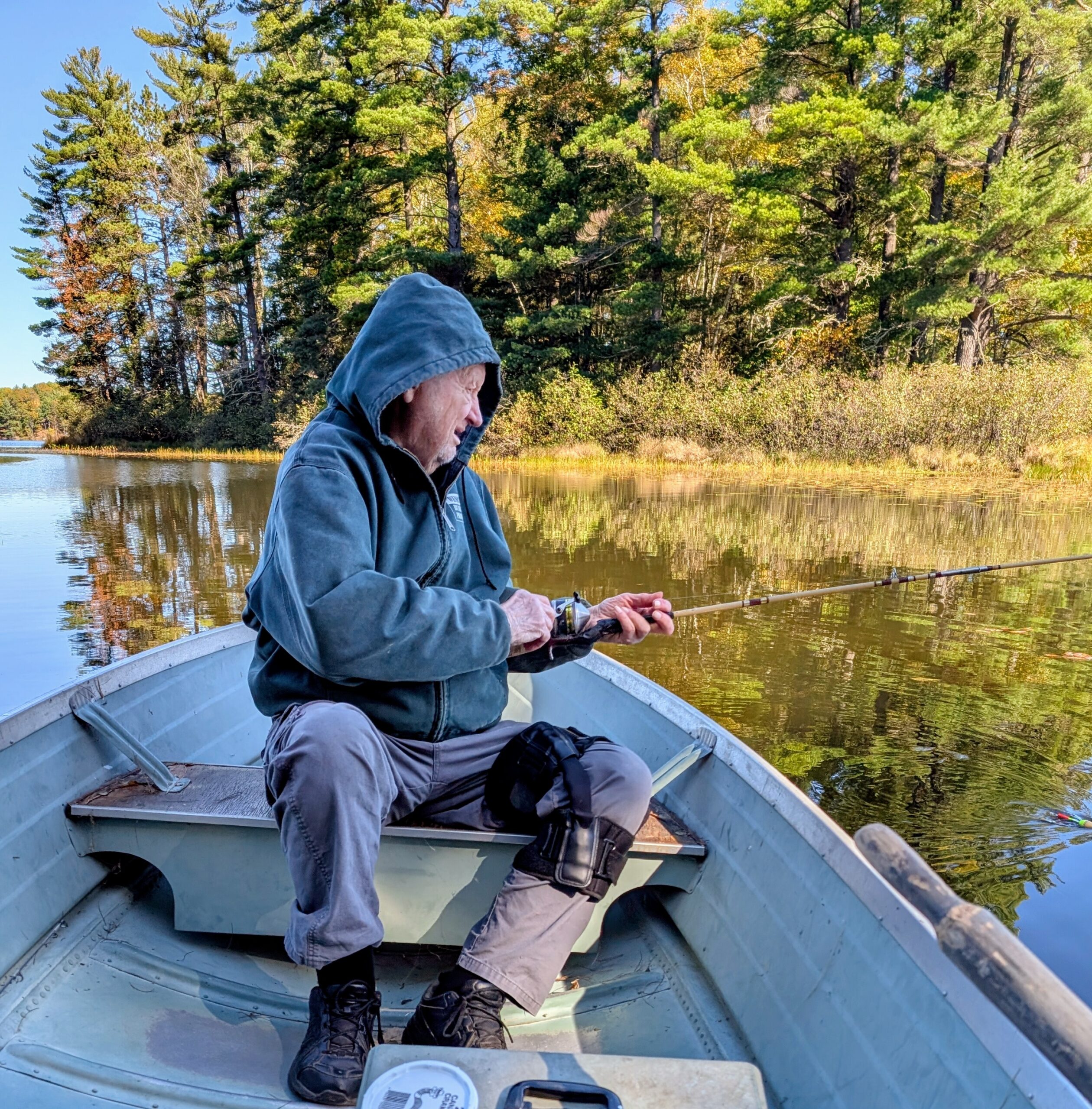 Person in a blue hoodie fishing from a small boat on a calm lake with trees in the background