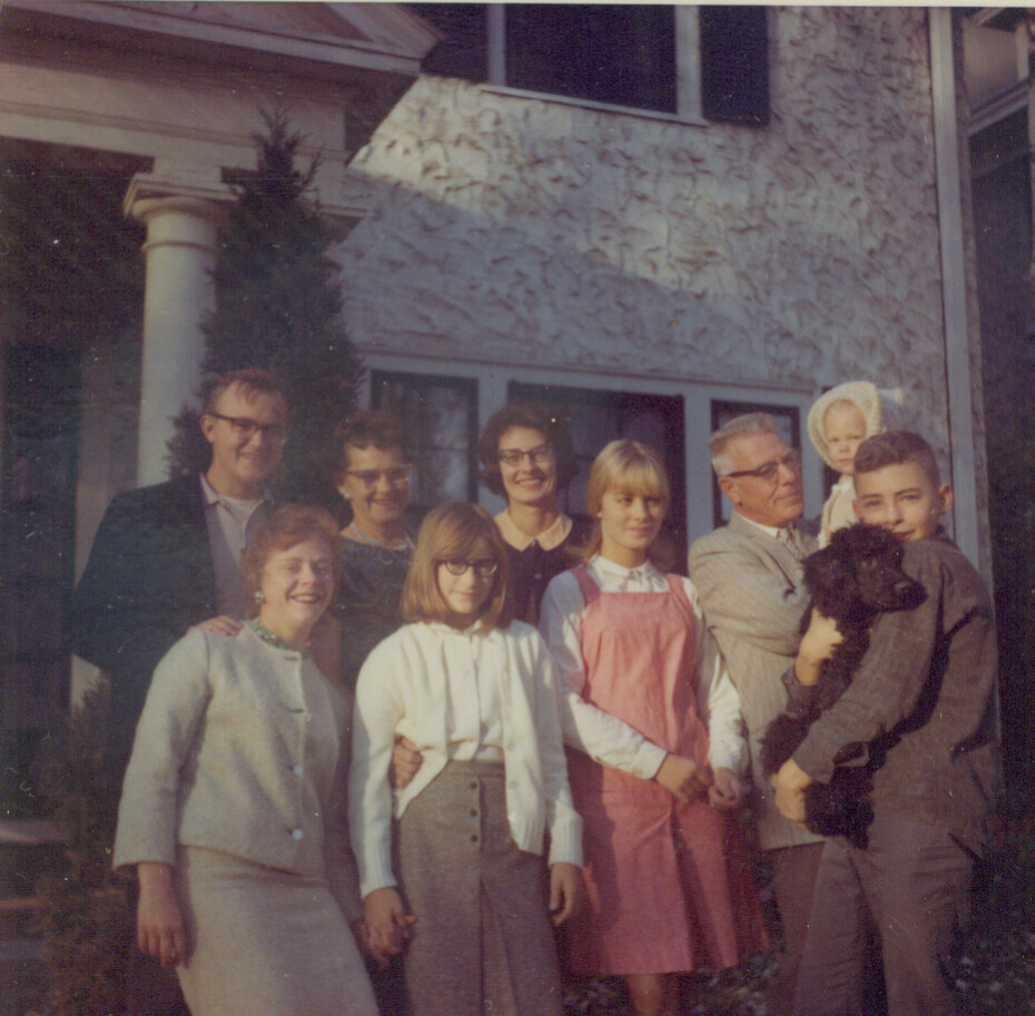 Multi-generational family posing on a front porch in front of a textured house, smiling at the camera; a man holds a small child, and another person holds a black poodle.