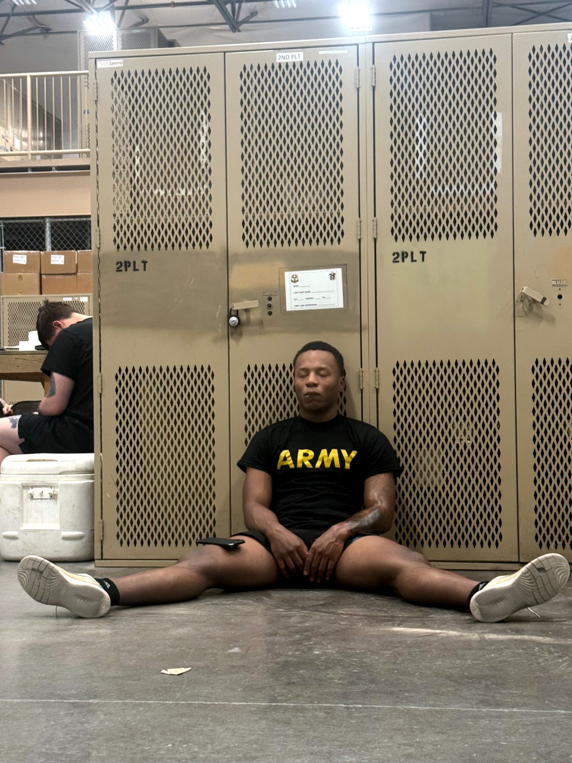 Man in a black 'ARMY' T-shirt sitting on the floor with legs spread in front of beige perforated lockers in an industrial space; another person sits to the left.
