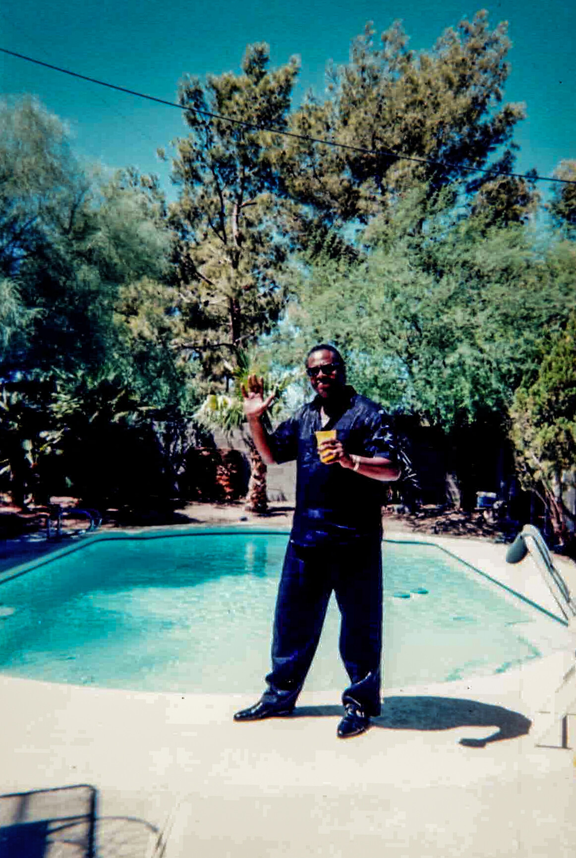 Man in a dark shirt and jeans stands by a swimming pool, waving and holding a yellow cup.