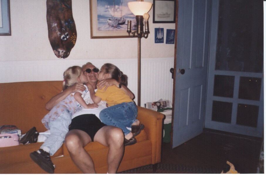 Woman on an orange sofa being kissed by three children in a cozy living room with a lamp and framed pictures on the wall.