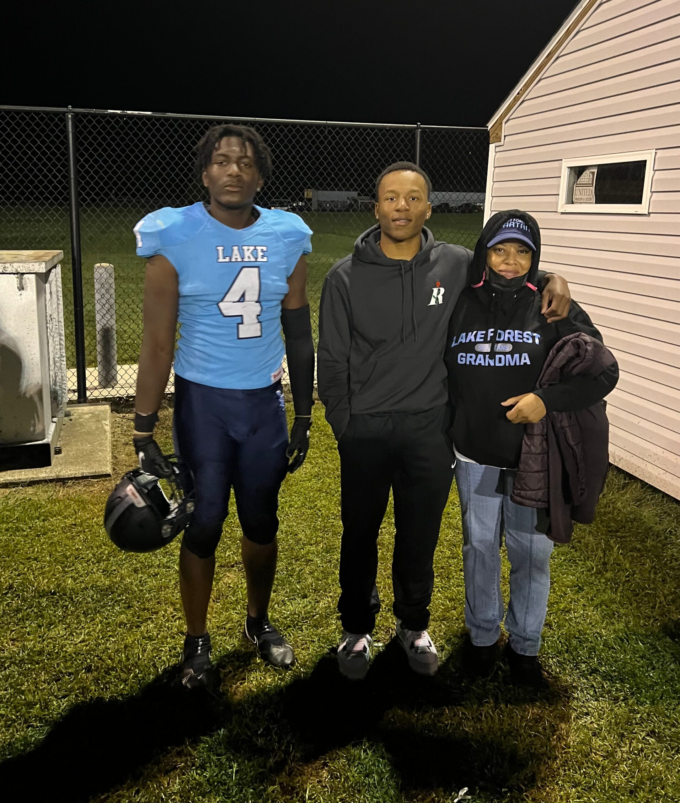 Three people pose together outdoors at night by a chain-link fence; two men in athletic wear and a woman in a Lake Forest Grandma sweatshirt stand close, smiling.