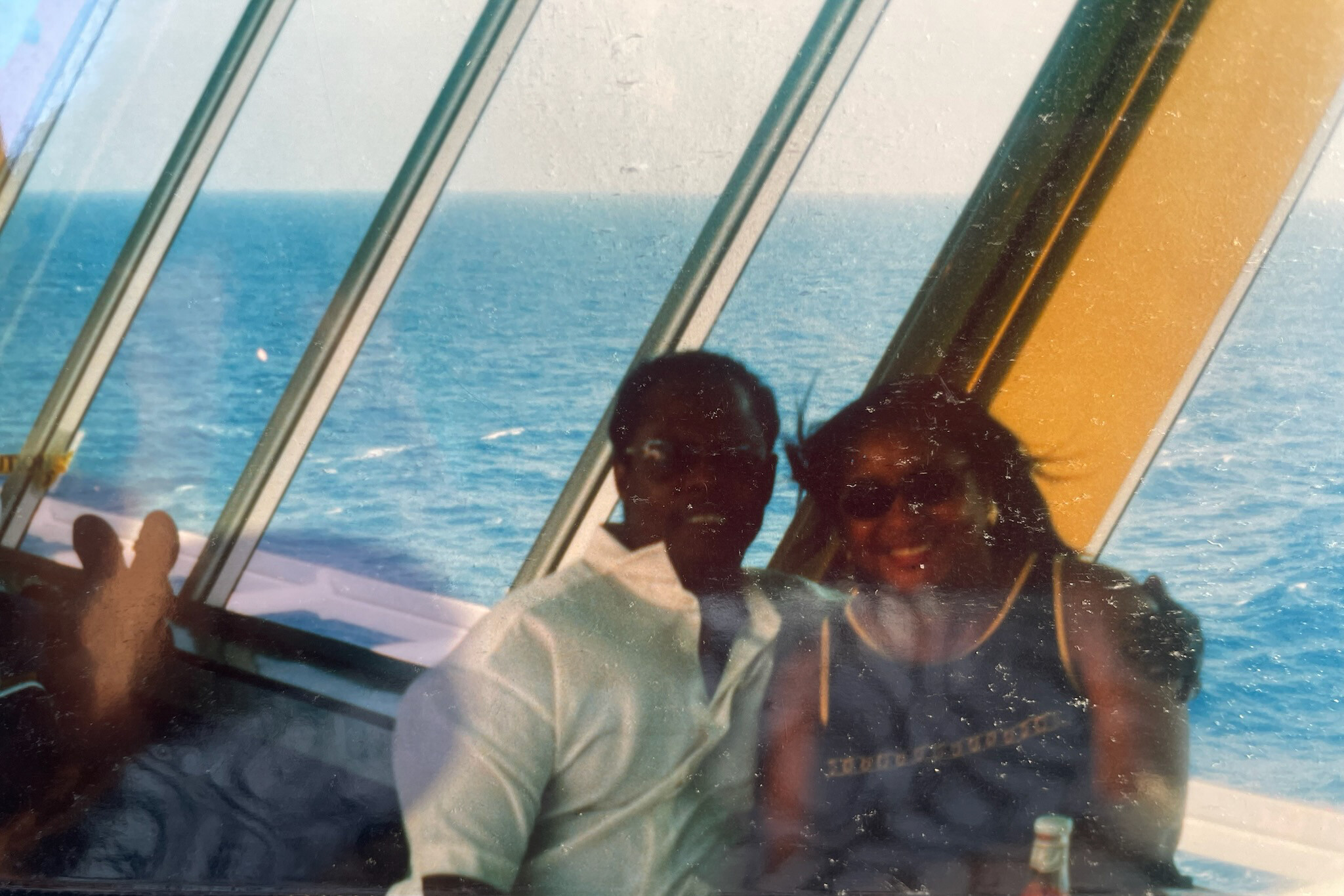 Two people appear as dark silhouettes reflected in a curved glass window, with the sea and sky behind them, giving a distorted view of a boat interior.