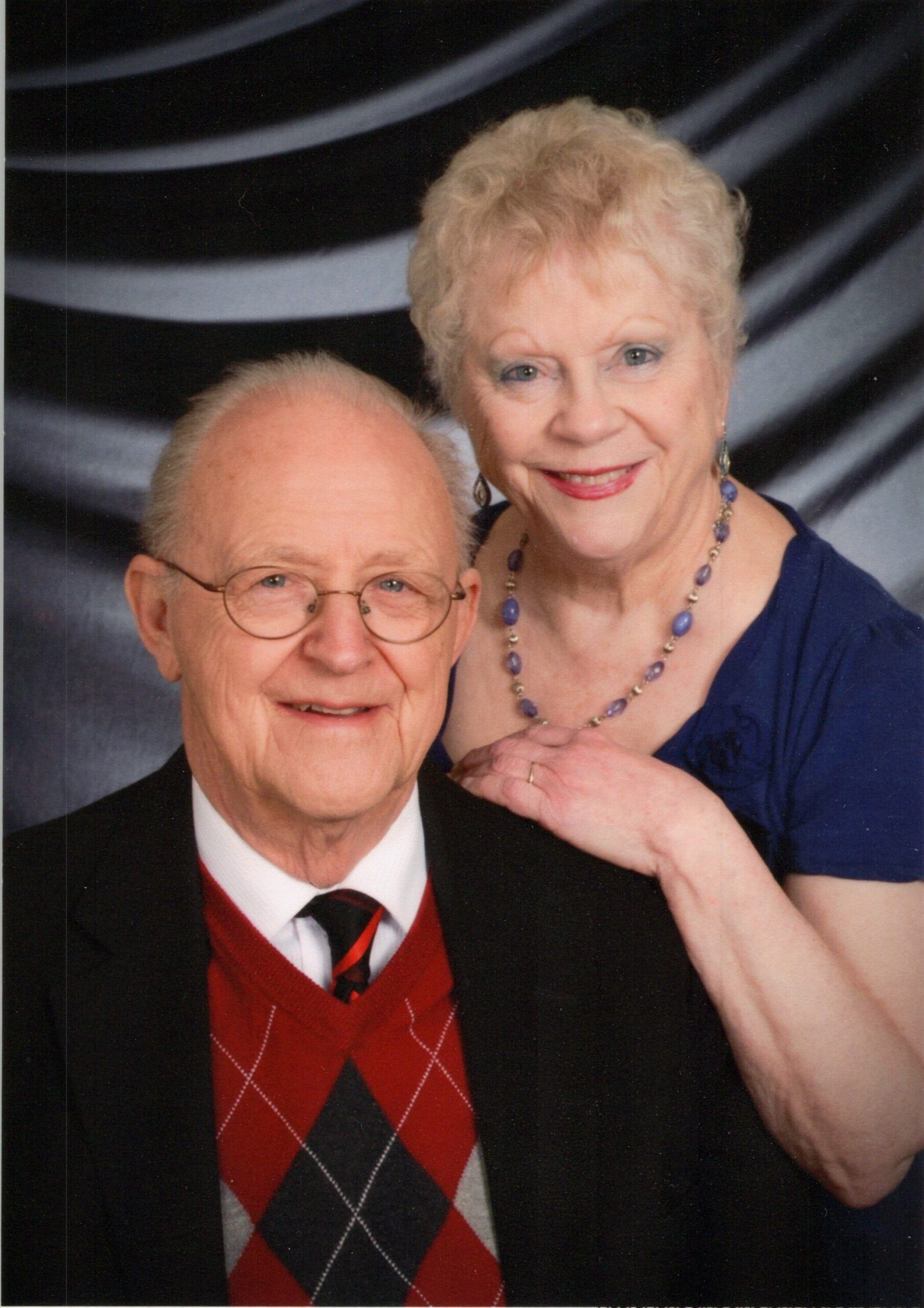 Elderly couple posing for a portrait: man in a suit with a red argyle sweater, woman in a blue dress and beaded necklace smiling behind him.
