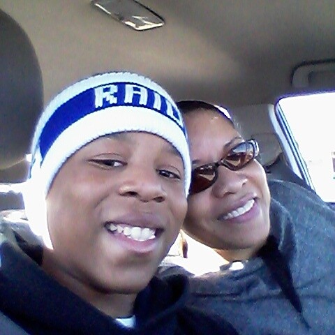 Two people smiling in a car; man in foreground wearing a white beanie with a blue stripe, woman beside him wearing sunglasses.