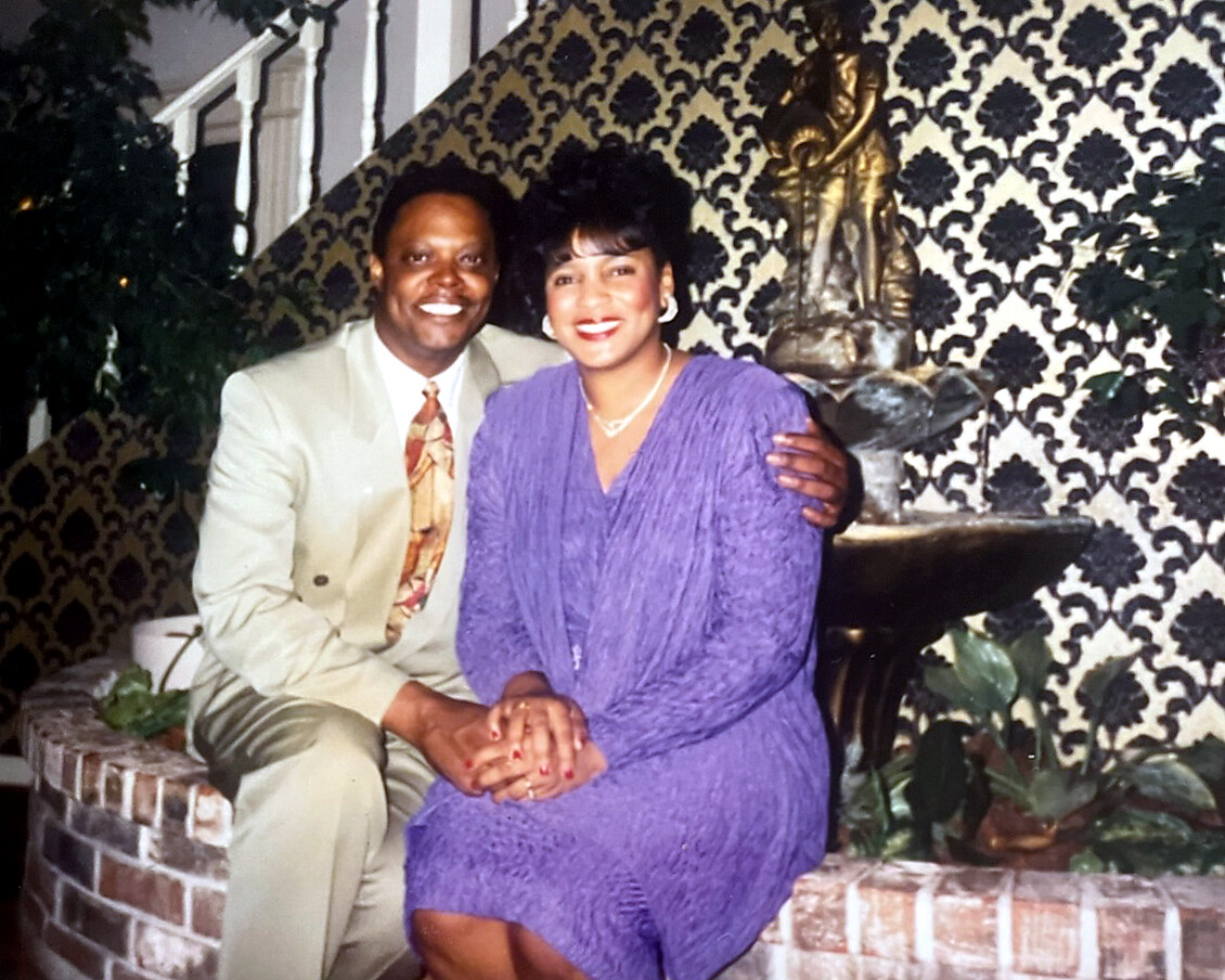 Smiling couple sitting close together on a brick planter, holding hands in front of a fountain and damask wallpaper backdrop.