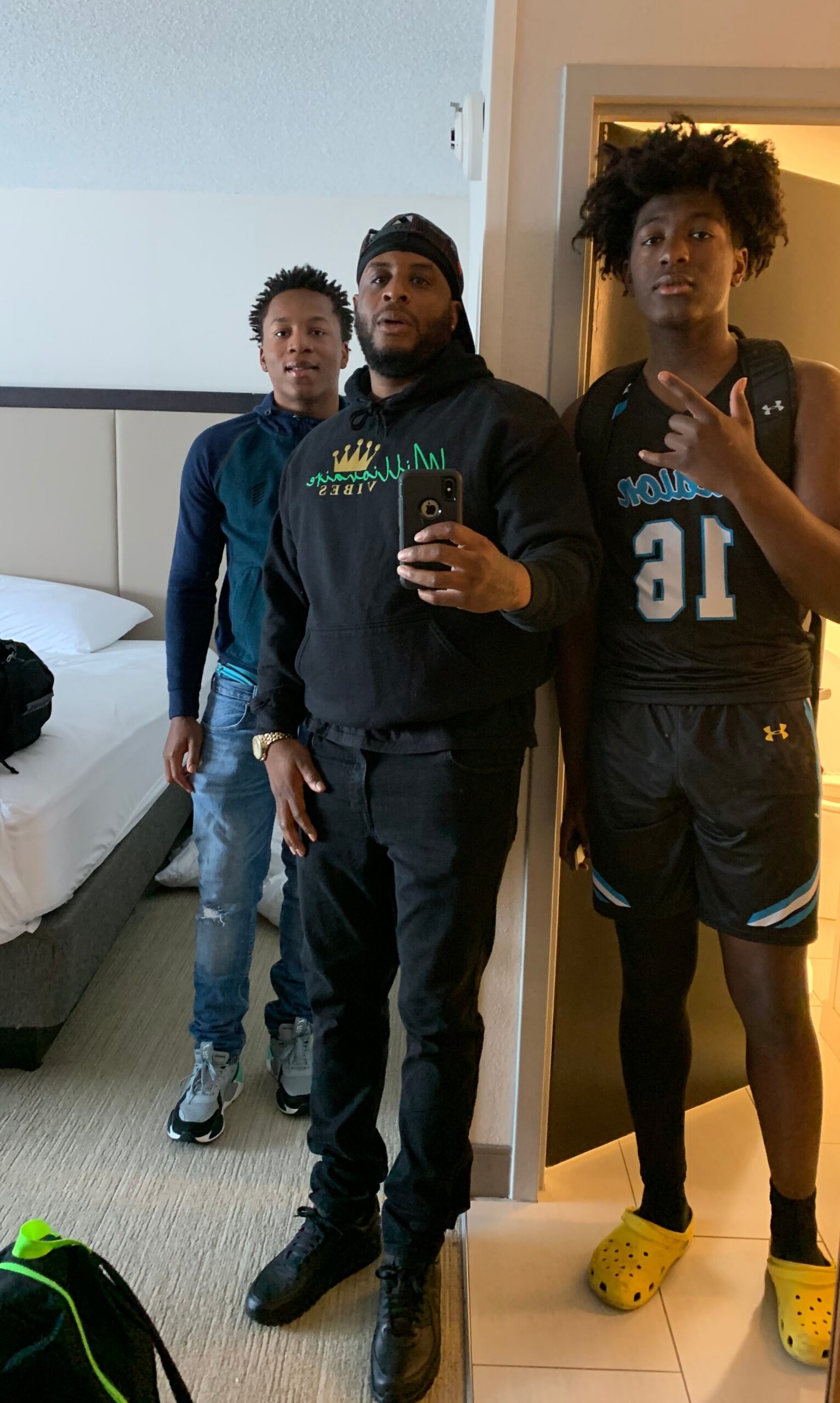 Three men taking a mirror selfie in a hotel room; center man in a black hoodie, others in casual athletic wear