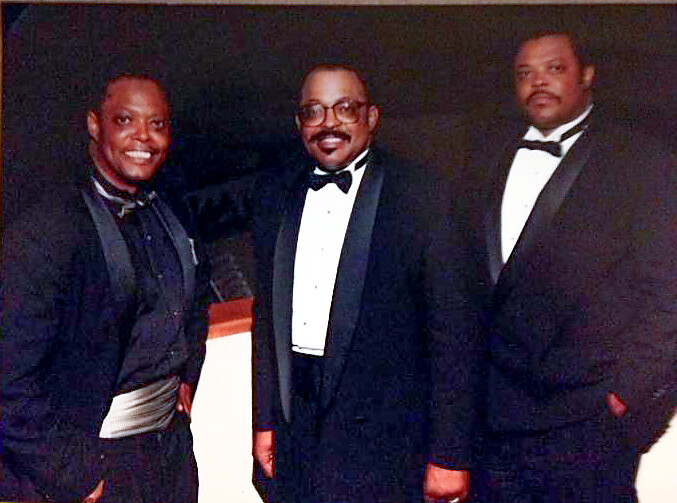 Three men in black tuxedos posing together at a formal event, smiling for the camera.