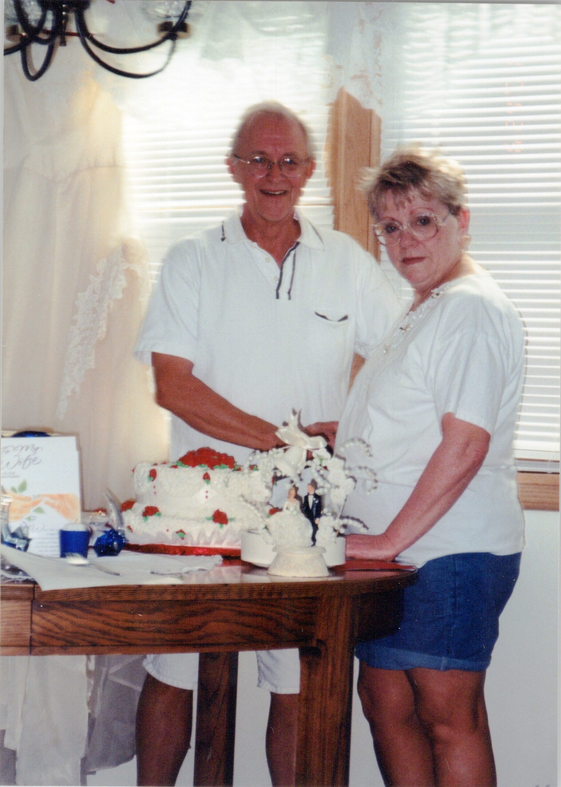 An elderly man and woman in white shirts cutting a white, flower-decorated cake at a celebration indoors.