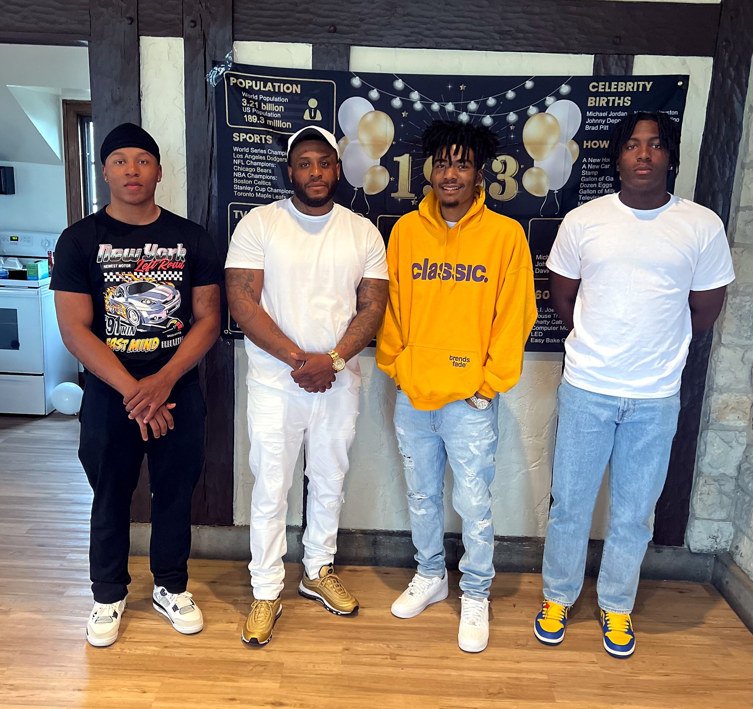Four men stand indoors shoulder to shoulder, dressed in casual streetwear and sneakers, in front of a dark backdrop with balloons and text.