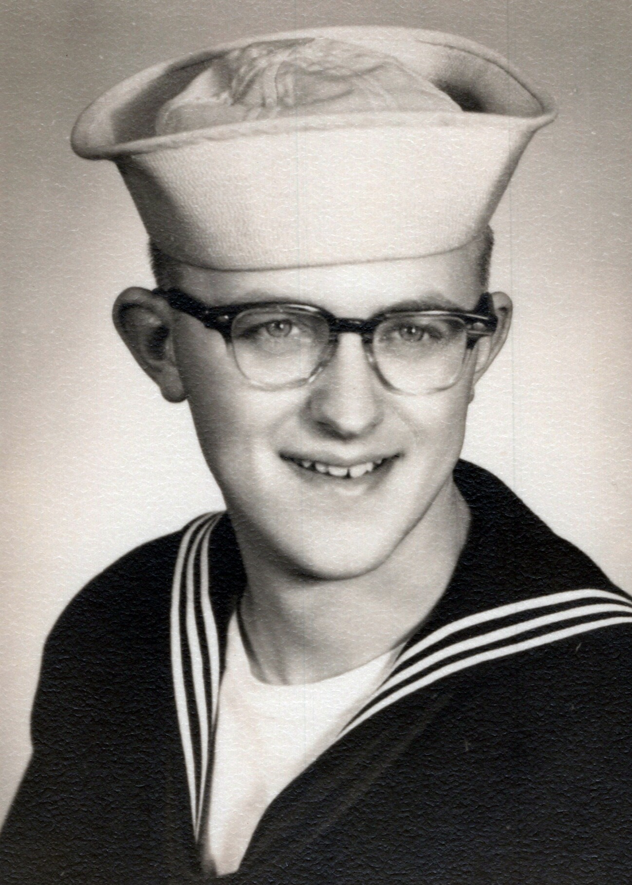 Young person in a vintage sailor uniform, white hat and glasses, smiling at the camera (black-and-white photo).
