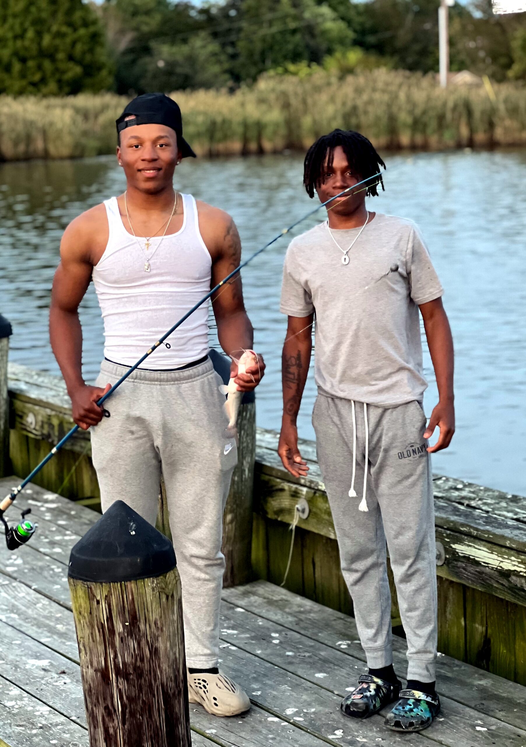 Two young men stand on a wooden dock by a lake, posing with fishing rods and a caught fish behind them in the water and reeds.