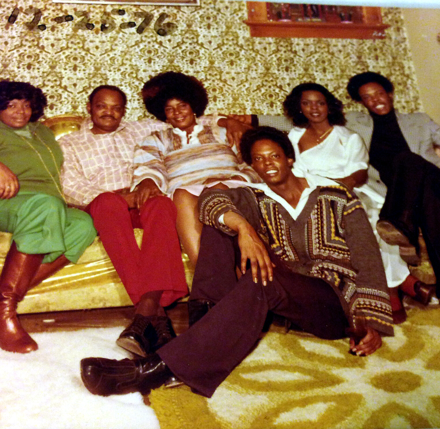 Six adults pose on a gold sofa in a 1970s living room with patterned wallpaper; they smile at the camera.