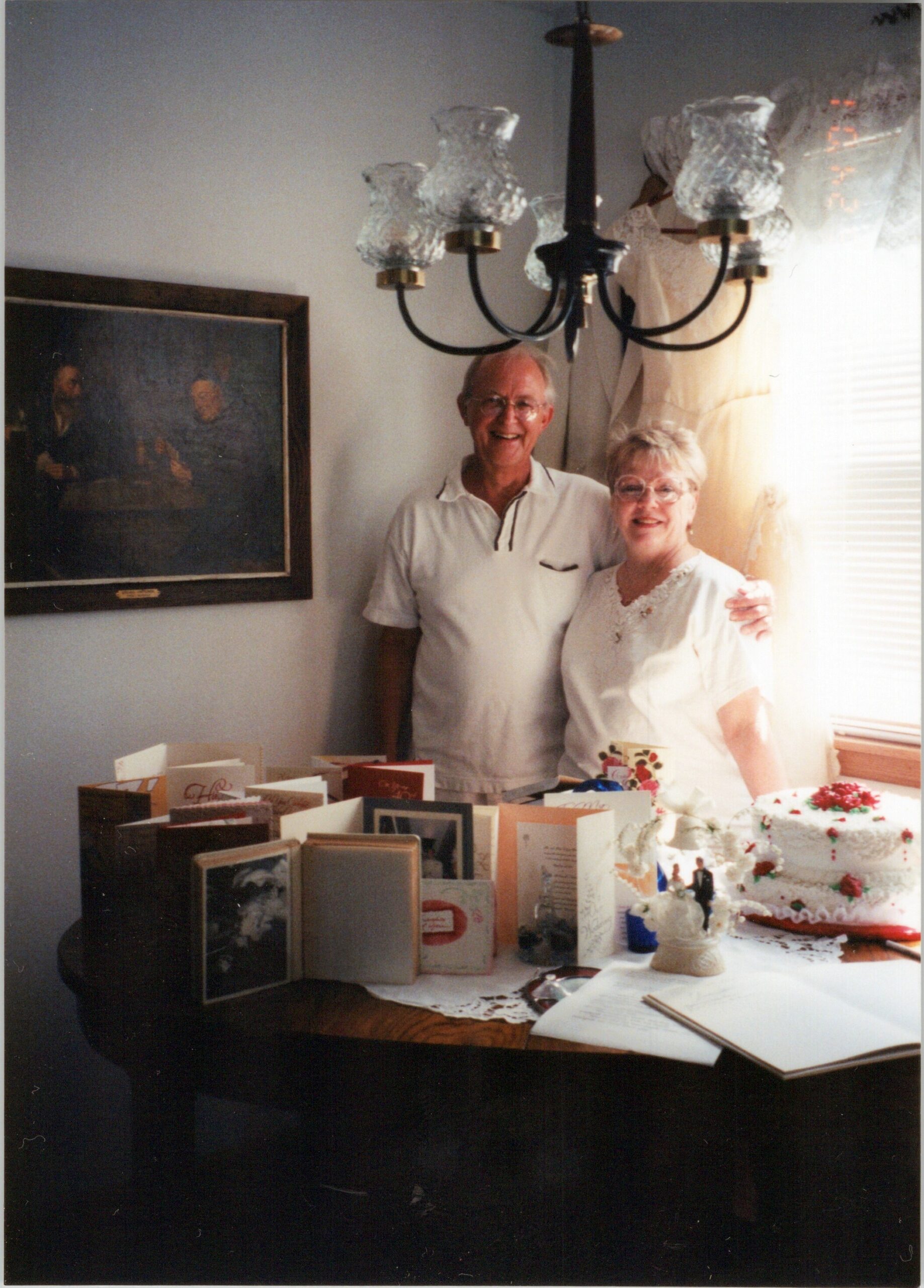 Older couple smiling and posing in a dining room with a cake and many cards on the table in front of them.
