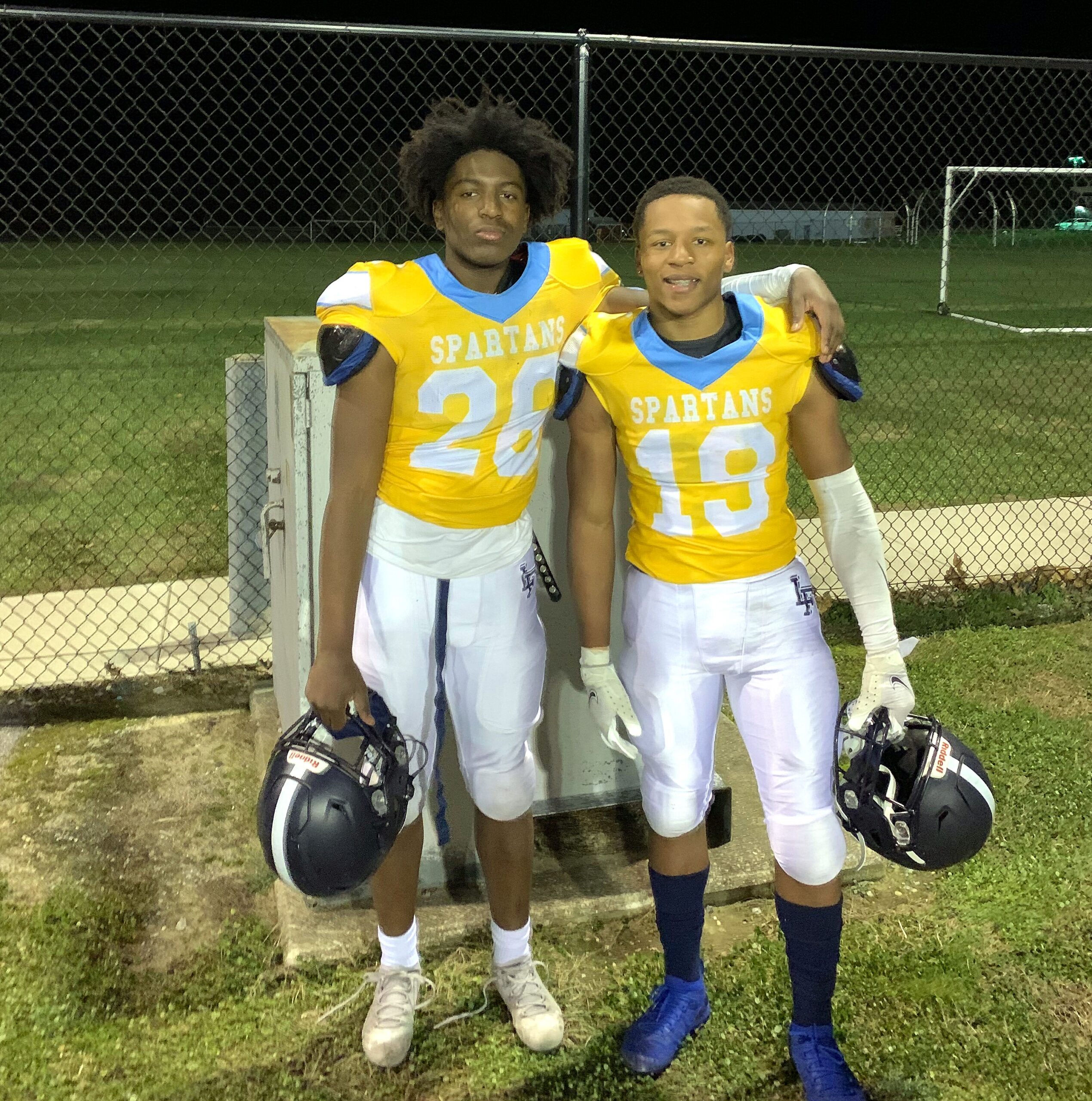 Two young football players in yellow Spartans jerseys pose together with helmets on the ground near a chain-link fence at night.