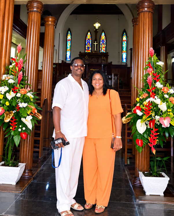 Couple posing in a church aisle between wooden columns, man with a camera and a woman in orange, with stained-glass windows behind them.
