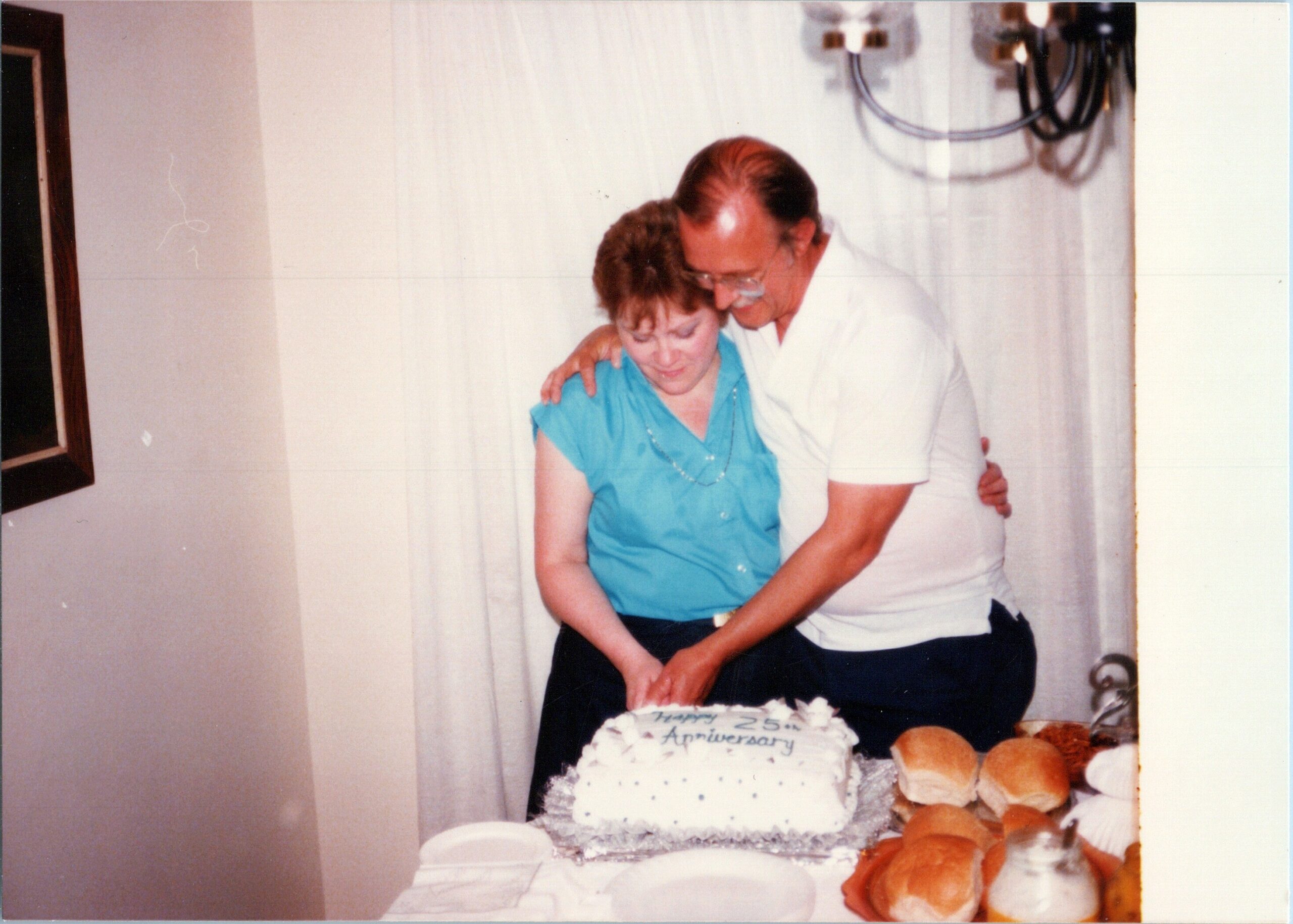 Older couple cutting a white anniversary cake labeled 'Happy 25th Anniversary' at a party, with bread rolls nearby on the table.