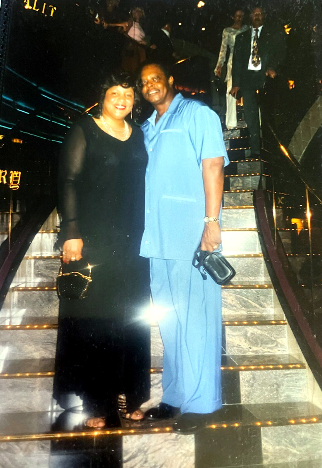 Smiling couple poses on a lit marble staircase in a nightclub; she wears a black dress, he in a blue outfit, posing together.