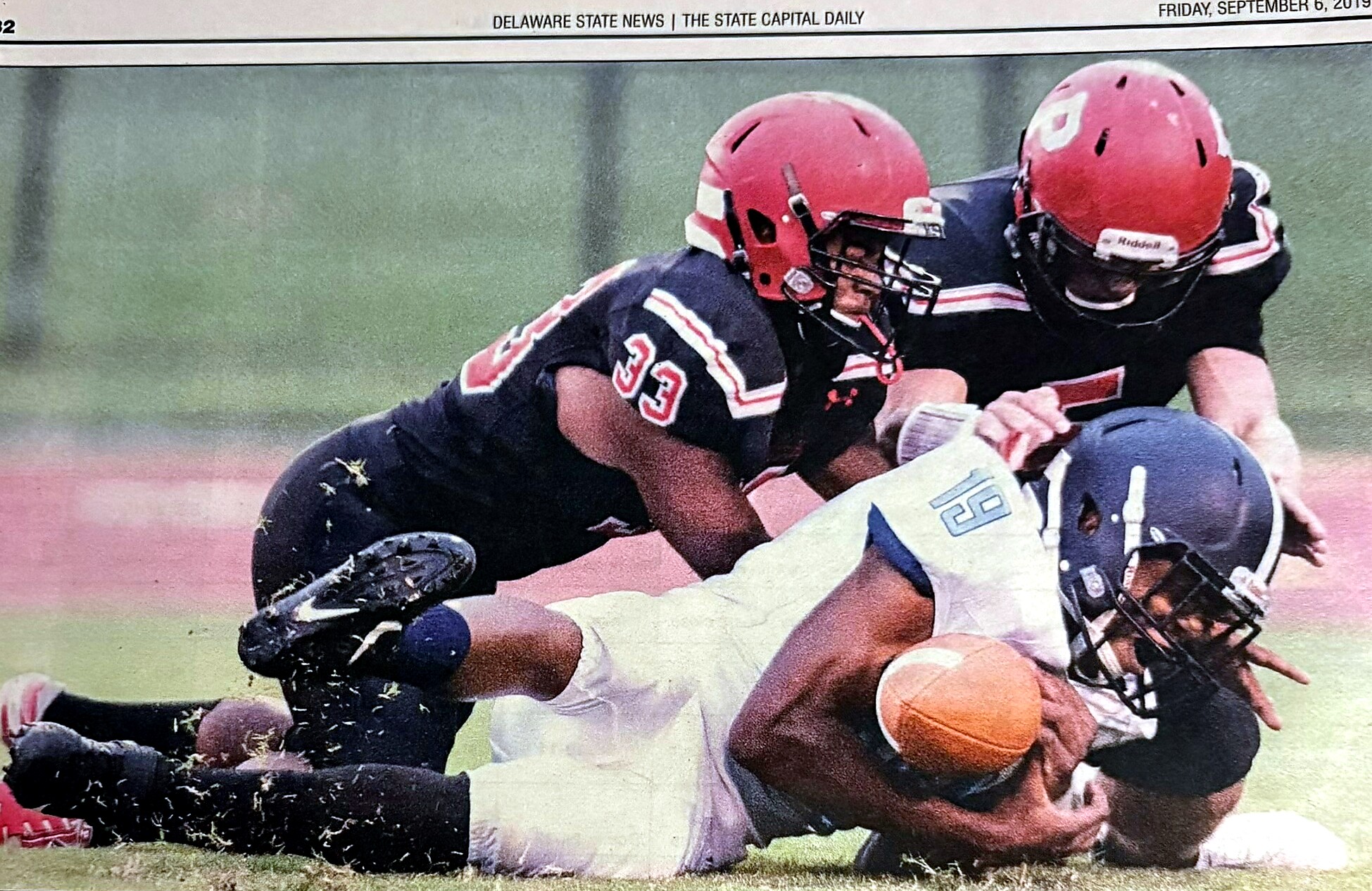 Two football players collide and bulk-tackle a ball carrier on a grassy field during a game; one wears a red helmet and black uniform with number 33, the other in white/blue.