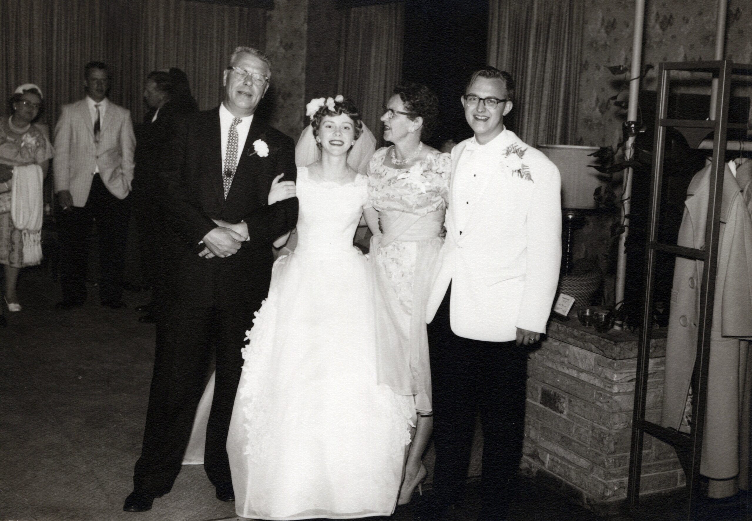 Bride in a white wedding dress and veil stands between an older man in a dark suit and a woman in a floral dress, with a smiling man in a white suit beside them at a reception.